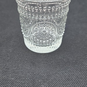 May include: A clear glass tumbler with an embossed floral and dot pattern. The glass has a thick base and a slightly flared rim. The design is symmetrical, with repeating patterns around the sides. The glass is likely used for drinking.