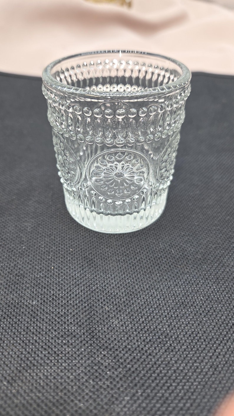 May include: A clear glass tumbler with an embossed floral and dot pattern. The glass has a thick base and a slightly flared rim. The design is symmetrical, with repeating patterns around the sides. The glass is likely used for drinking.