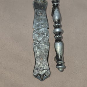 May include: Two antique metal cabinet hardware pieces. One is a long, textured plate with a decorative top and bottom. The other is a handle with a detailed, rounded design. Both have a silver finish.