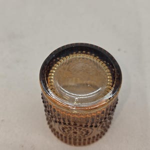 May include: A small, round, amber-colored glass container with a decorative pattern of raised dots and floral designs. The rim is a darker shade of brown. The container is likely a small jar or votive holder.