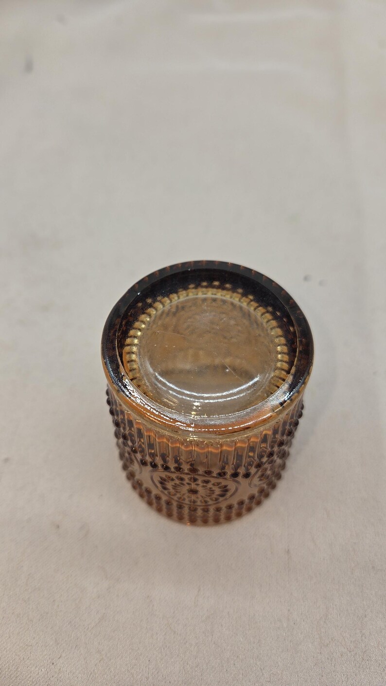 May include: A small, round, amber-colored glass container with a decorative pattern of raised dots and floral designs. The rim is a darker shade of brown. The container is likely a small jar or votive holder.