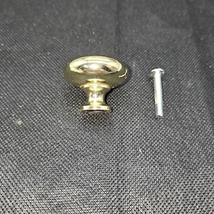 May include: A gold-colored cabinet knob with an oval top and a decorative base. A silver-colored screw is next to the knob. The knob is likely for furniture such as drawers or cabinets.