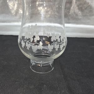 May include: Clear glass candle holder with a flared top and a base. The glass is decorated with a white clover pattern around the middle. The candle holder is designed to hold a small candle.