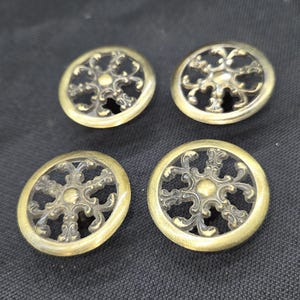 May include: Four round metal buttons with an antique brass finish. Each button features a circular frame surrounding an intricate, openwork filigree design with a central raised dot and radiating scroll patterns throughout the interior.