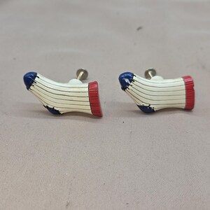 May include: Two decorative cabinet knobs shaped like baseball socks. Each knob features a white sock with black stripes, a red cuff, and a blue toe. The knobs have a metal screw for attachment.