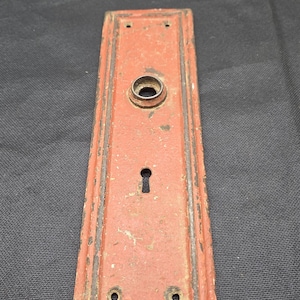 May include: A rectangular metal door plate with a weathered red finish and dark edges. The plate features a central circular opening for a knob, a keyhole below it, and four small screw holes located at the top and bottom corners of the piece.