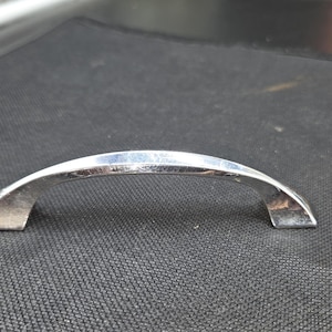 May include: A polished chrome cabinet handle with a smooth, arched design. The hardware features a reflective metallic finish and two mounting points at the ends, showcasing a curved, elongated shape that is consistent in its silver-toned appearance.