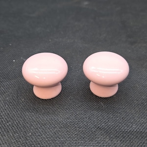May include: Two matching cabinet knobs with a smooth, glossy, light pink finish. Each knob features a rounded, mushroom-shaped top and a tapered base, designed for attachment to furniture drawers or cabinet doors for opening and closing.