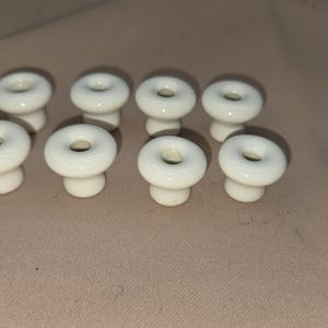 May include: Eight white ceramic cabinet knobs with a donut-shaped design. The knobs have a glossy finish and a small hole in the center. They are arranged on a neutral-colored surface, suitable for furniture restoration or DIY projects.