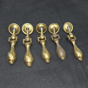 May include: Five antique brass drawer pulls with a teardrop design. Each pull features a circular backplate, a rectangular connector, and a decorative, elongated handle. The pulls are arranged in a row against a dark background.