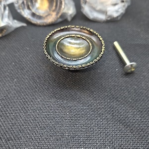 May include: A round cabinet knob with a dark, iridescent finish and a textured, antiqued gold-toned rim. The knob features a central concave depression surrounded by a beaded border. A single metal mounting screw is placed beside the knob.