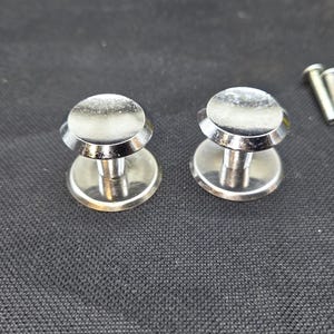 May include: Two silver-colored metal buttons with a smooth, rounded top surface and a cylindrical base. The buttons feature a polished, reflective finish and a distinct flared edge design. A small metal screw is visible to the right of the buttons.