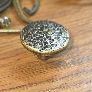 May include: A round cabinet knob with a textured, hammered surface in a dark antique brass finish. The knob features a raised, uneven top with deep crevices and a metallic sheen, accompanied by a metal mounting screw extending from the back.