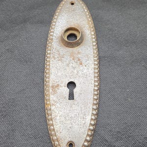 May include: An oval-shaped metal door escutcheon plate with a weathered, textured finish. The plate features a beaded border, a central circular opening for a door knob, a lower keyhole opening, and two small screw holes at the top and bottom.