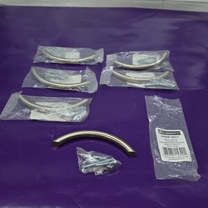 May include: A collection of brushed nickel cabinet pulls, each in a curved, half-moon shape. Several are packaged in clear plastic, with mounting screws visible. One pull and a packet of screws are loose. A product label is visible.