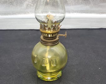 May include: A vintage oil lamp with a clear glass chimney and a green-tinted glass base. The lamp features a brass burner and a small handle for adjusting the wick. The base has a rounded shape with embossed details.