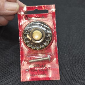 May include: A decorative cabinet knob with a bronze finish, featuring a floral design around the perimeter and a central circular accent. Includes a screw for installation, packaged in a red and clear plastic blister pack.