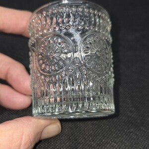 May include: Clear glass votive candle holder with an embossed floral and geometric pattern. The cylindrical holder is held by a hand against a dark background. The glass has a detailed, textured surface.