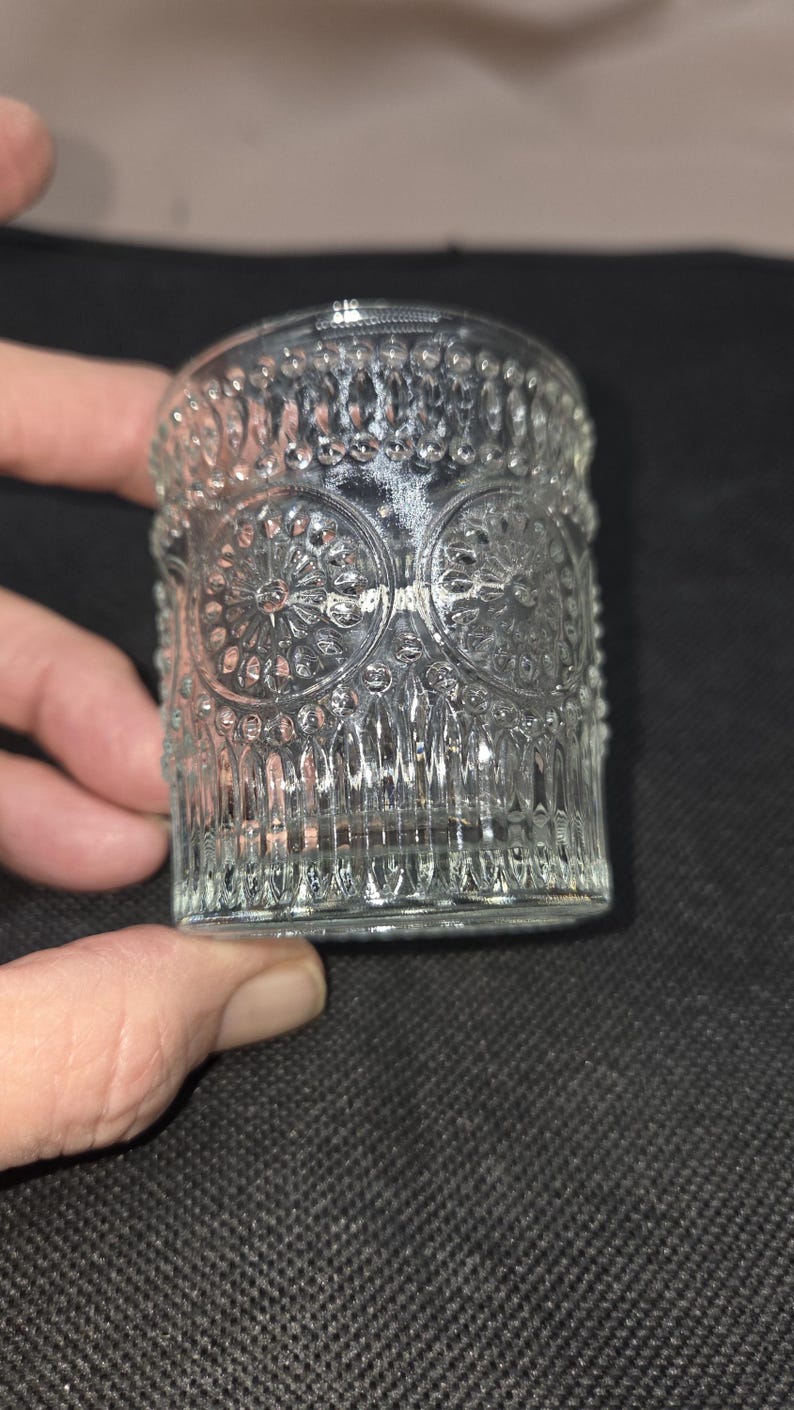 May include: Clear glass votive candle holder with an embossed floral and geometric pattern. The cylindrical holder is held by a hand against a dark background. The glass has a detailed, textured surface.