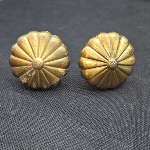 May include: Two antique brass-colored drawer pulls with a floral design. Each pull has a central circular element surrounded by radiating petals. The pulls are set against a dark gray textured background.