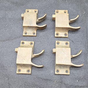May include: Four identical brass window sash locks arranged in a two-by-two grid. Each hardware piece features a rectangular mounting plate with four screw holes and a curved, protruding lever arm designed for securing a window sash in place.