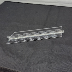 May include: A clear, plastic sign holder with a rectangular shape and a small lip on one end. The holder has multiple parallel lines etched into its surface, creating a textured appearance. It is designed to hold a sign or label.