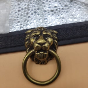 May include: A brass-toned metal furniture pull featuring a detailed lion head design with a circular ring handle hanging from the lion's mouth. The hardware includes a single threaded bolt for attachment, showcasing a weathered metallic finish.