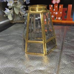 May include: A clear glass lampshade with a brass-colored metal frame. The lampshade has a geometric, hexagonal shape. The top and bottom are wider than the middle section. The lampshade is approximately 6 inches tall.