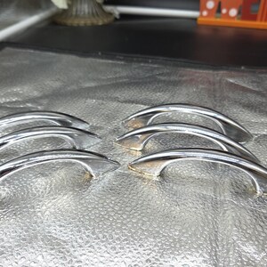 May include: A set of six polished chrome cabinet pulls arranged in two rows of three. Each handle features a smooth, arched design with tapered ends and a reflective metallic finish, designed for attachment to furniture or cabinetry doors.