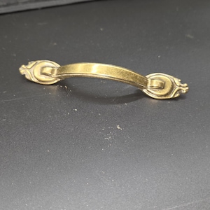 May include: A brass-colored cabinet pull with a curved handle and decorative ends. The hardware is designed with a classic style, suitable for furniture restoration or new builds. The pull is shown against a dark background.