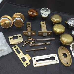 May include: A collection of vintage door hardware including various round door knobs in brass, chrome, and glass finishes, along with rectangular and oval metal backplates, latch mechanisms, metal spindles, and a small bag of assorted mounting screws.