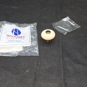 May include: A round, cream-colored cabinet knob with a metallic base, accompanied by a small plastic bag containing a metal mounting screw and a larger plastic bag featuring the Hickory Hardware logo and company information printed in blue and red.
