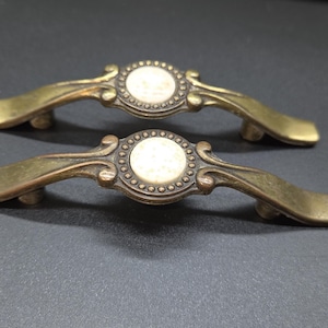 May include: Two antique brass drawer pulls with a curved handle design. Each pull features a central circular ivory-colored insert surrounded by a decorative border. The pulls are set against a dark background.