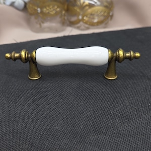 May include: A cabinet pull with a white ceramic center and antique brass-colored metal ends. The handle is a curved, elongated shape, with decorative detailing on the ends. The hardware is designed for furniture and home decor.