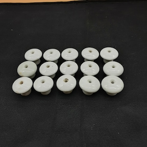 May include: A collection of 15 white ceramic insulators, each with a small hole in the center. The insulators are arranged in a staggered pattern on a black surface.