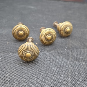 May include: Four round cabinet knobs made of antique brass-toned metal. Each knob features a central circular protrusion surrounded by a series of radiating, embossed lines that extend to the outer edge, creating a sunburst-like pattern.