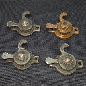 May include: Four vintage metal window sash locks arranged in a two-by-two grid. Each fastener features a circular base with a rotating lever and a curved hook. The hardware displays varying degrees of oxidation, including dark grey, rusted brown, and muted green tones, showing signs of age and wear on the metallic surfaces.