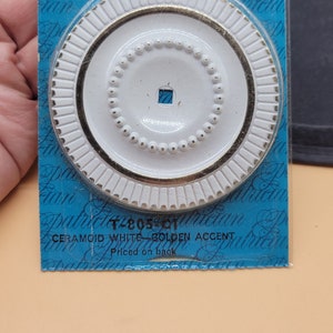 May include: A white ceramic decorative accent with a gold trim and a ring of small gold beads around the center. The accent is packaged on a blue background with the text "Amerock Ceramoid White-Golden Accent T280581 Priced on back".