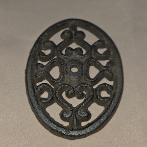 May include: A dark gray, cast iron trivet with an oval shape. The trivet features an openwork design of swirling, symmetrical patterns. The metal has a slightly textured appearance. This decorative item is designed to protect surfaces from hot items.