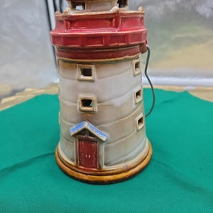 May include: A ceramic lighthouse candle holder with a blue top, red roof, and white body. The lighthouse has small square windows and a small door. A metal handle is attached to the side. The base is a golden yellow color.