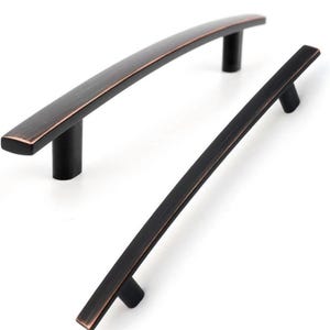 May include: Two bronze-finished cabinet pulls. One is curved and larger, the other is straight and smaller. Both have a rectangular bar and cylindrical posts. The hardware is set against a white background.