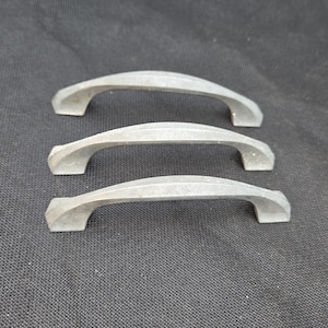 May include: Three silver-toned metal cabinet handles arranged in a vertical stack. Each handle features a curved, arched design with a central ridge running along the top surface and flat mounting points at both ends for attachment to furniture.
