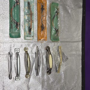 May include: A collection of cabinet hardware, including handles and pulls, in various styles and finishes. The hardware is packaged in clear plastic and cardboard. Finishes include copper, silver, gold, and bronze.