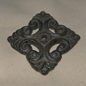 May include: Ornate, dark gray metal decorative piece with a diamond shape and intricate scrollwork design. The center features a small circular opening. This decorative element could be used for furniture or architectural accents.