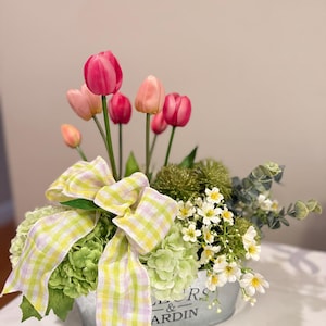 May include: A floral arrangement featuring pink tulips, green hydrangeas, and small white flowers in a galvanized metal container. A yellow and white gingham bow adds a decorative touch. The container has the text "Flowers & Garden".