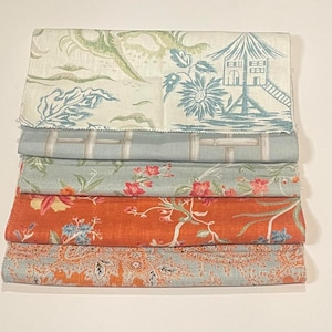 May include: A stack of five fabric swatches with different patterns. The top swatch is white with a blue and green floral pattern and a blue pagoda design. The second swatch is light blue with a white geometric pattern. The third swatch is light blue with a floral pattern in shades of pink and green. The fourth swatch is orange with a floral pattern in shades of orange and green. The bottom swatch is light blue with a paisley pattern in shades of orange and blue.