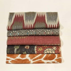 May include: A stack of five fabric swatches with different patterns and colors. The top swatch is a red, gray, and white ikat pattern. The second swatch is a brown and blue geometric pattern. The third swatch is a red and white floral pattern. The fourth swatch is a dark brown and green floral pattern. The bottom swatch is a brown and white animal print.