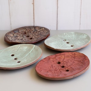 May include: Four ceramic soap dishes in shades of brown, red, and green. Each oval dish has a leaf or floral pattern and three drainage holes. The dishes are arranged on a gray surface against a white wooden background.