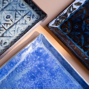 May include: Overhead shot of four square ceramic plates in shades of blue. Each plate features a unique geometric pattern and texture. The plates are arranged on a neutral background, showcasing their intricate designs and colors.