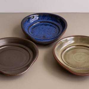 May include: Three handmade ceramic dishes in shades of blue, brown, and green. Each dish has a unique shape with a slightly raised rim and a smooth, glossy finish. The dishes are arranged on a neutral surface.